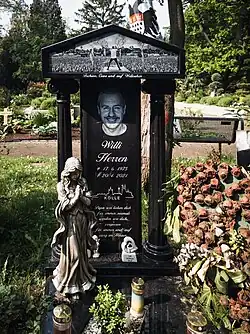 Grabstein aus schwarz-glänzendem Material, zentrale Platte mit 2 seitlichen dorischen Säulen, die einen schlichten Giebel tragen. Platte: gefrästes Porträt Willi Herren, darunter Lebensdaten, Kölner Skyline mit „Kölle“, darunter in Schreibschrift: Papa wir lieben dich, niemals werden wir dich vergessen. Für immer und ewig im Herzen. Giebel: vollflächig gefrästes Bild, das einen Künstler von hinten auf der Bühne mit erhobenen Armen und Blick ins Publikum zeigt. Vor dem Grabstein: große Engelfigur und diverse Deko.