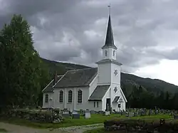 Foto einer weißen Holzkirche, dahinter steigt die Landschaft an