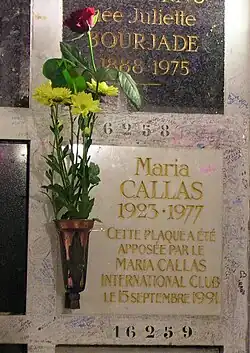 Maria Callas Gedenkplatte des Urnengrabes mit der Aufschrift 'Maria Callas 1923 1977 Cette Plaque a Ete Aposee Par Le Maria Callas International Club Le 15 September 1991. Eine kleine metallernde Vase befüllt mit 4 gelben Blumen und einer Rose ist mit einem dunkelmetallenden Fassung befestigt. Unter der Platte befindet sich die Nummer 16259 darüber 16258 mit einem weiteren Namen. Rings um dem Grab sind zahlreiche Kritzeleien und Unterschriften mit Herzchen in verschiedenen Farben zu erkennen.