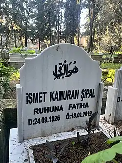 Das Bild zeigt einen Grabstein auf einem Friedhof. Der Grabstein ist aus weißem Marmor gefertigt und trägt Inschriften in Arabisch und Türkisch. Die arabische Inschrift oben lautet „هو الباقي“, was ins Deutsche übersetzt „Er ist der Ewige“ bedeutet. Darunter steht die türkische Inschrift: İSMET KAMURAN ŞİPAL RUHUNA FATİHA D: 24.09.1926 Ö: 18.09.2019 Die Übersetzung lautet: İSMET KAMURAN ŞİPAL Fatiha für die Seele Geboren: 24.09.1926 Gestorben: 18.09.2019 Der Grabstein befindet sich in einer von Bäumen gesäumten, grünen Umgebung. Im Hintergrund sind weitere Grabsteine sowie hohe, schattenspendende Bäume zu sehen. Das Bild vermittelt eine ruhige, friedliche Atmosphäre.