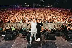 das Duo steht nachts auf der Bühne mit Publikum im Hintergrund. Einer der beiden streckt die Faust mit einem Mikrofon in die Höhe