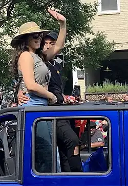 Hailie Sahar steht in einem dunkelblauen Auto mit offenem Dach und winkt der am Rand stehenden Menge zu. Sie trägt ein graues Top, eine Jeans, eine schwarze Sonnenbrille und einen beigenen Sonnenhut mit zwei gelben Streifen in der Mitte. Neben ihr steht ein Mann mit schwarzer Kappe, schwarzem T-Shirt und schwarzer Hose. Er hat einen regenbogenfarbenen Herz-Anstecker an der linken Brust und hält seine Hand auf Sahars Hüfte. Im Hintergrund sind ein Baum mit dunkelgrünen Blättern, ein beiges Wohnhaus, ein Balkon mit spitzen, hellgrünen Pflanzen und Passanten verschiedenen Alters zu sehen.