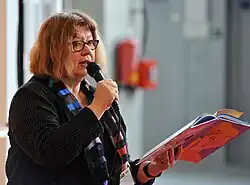 Das Farbfoto zeigt Hannele Huovi ein Mikrofon in der einen Hand und ein Buch der anderen Hand haltend bei einer Vorlesung im Jahr 2011