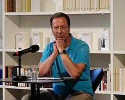 Pleschinski während einer Lesung an Tisch mit Mikro, Hände unters Kinn gestützt