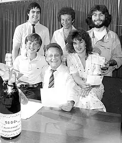 Hans Rosenthal mit „Sonntagsrätselkindern“ anlässlich der 1000. Sendung. Das Bild zeigt eine Gruppe von sechs Personen, die um einen Tisch versammelt sind. Hans Rosenthal im Anzug hält in der Hand ein Schreiben, während die anderen lächeln und Postkarten zeigen. Auf dem Tisch befindet sich eine große Champagnerflasche mit einer Schleife um den Hals. Auf der Flasche steht der Text „WUNSCH 1000x KLINGENDES SONNTAGSRÄTSEL“.Im Hintergrund ist ein Vorhang sichtbar, der den Ort der Veranstaltung gemütlich wirken lässt. Die Personen tragen eine Mischung aus Freizeit- und halbformeller Kleidung.