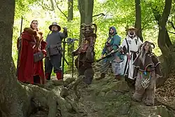 Auf einem Felsabsatz in einem lichten Wald hat sich eine Gruppe aus sechs Personen positioniert, die vermutlich einen Magier, einen Söldner, eine Art Minotaurus, eine Zwergin, einen Priester und einen Faun darstellen.