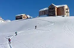 zwei große aus Holz erbaute Berghütte mit Schnee auf dem Dach auf einem Bergplateau, auf dem schneebedeckten Hang unter ihnen steigen Skiwanderer in einer S-Kurve auf