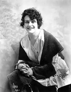 Schwarz-Weiß-Porträtfoto einer lächelnden, sitzenden Helen Gibson vor hellem Hintergrund. Sie trägt einen dunklen Mantel und ein weißes, ungefähr bis zu den Knien reichendes Oberteil. Ihre dunklen Haare trägt sie als lockigen, bis zu den Ohren reichenden Rundschnitt. Ihre Hände liegen mit ineinander verschlungenen Fingern im Schoß. In der linken Fotoecke befindet sich die weiß geschriebene Signatur des Fotografen.