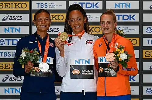 Anna Hall im blauem Trainingsanzug, Katarina Johnson-Thompson in weißer Trainingsjacke und Anouk Vetter in Orange präsentieren ihre Medaillen vor einer Sponsorentafel.