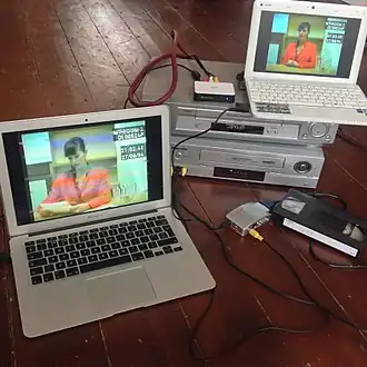 Ein Laptop mit den Aufnahmen des Spiels steht auf zwei VHS-Rekordern. Drähte verbinden sie zu einem naheliegenden Laptop, connect them to another laptop nearby, mit einer VHS-Kartusche auf der rechten Seite.