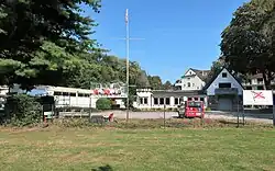 Hinter einem Rasenstück und einem Zaun liegt ein Parkplatz. Dahinter sind ein Bootshaus und ein Vereinslokal zu erkennen.