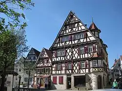 Um einen Platz angeordnet: eine Stele mit der Aufschrift „Place de Tarare“, Fachwerkhäuser und ein Brunnen