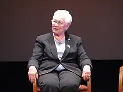 Sitzende Frau mit kurzen weißen Haaren, ihr Körper ist dem Fotografen zugewandt, sie blickt leicht lächelnd zu Seite. Sie trägt einen dunkelgrauen Hosenanzug.