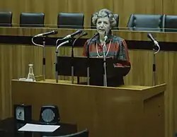 Eine Frau mit kurzer grauer Föhnfrisur steht an einem Pult hinter mehreren Mikrofonen. Sie trägt eine Bluse mit großem blau-rotem Karo-Muster. Auf dem Pult steht eine Karaffe mit Wasser. Auf einem Tisch vor dem Pult stehen eine große analoge Uhr und ein Lautsprecher. An der Qualität des Bildes und dem Design der Mikrofone, Lautsprecher, und der Frisur der Person ist ableitbar, dass das Bild aus den 1980er Jahren stammt.