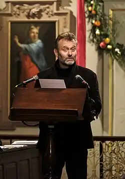 Hugh Dennis bei einer Rede im Namen des National Churches Trust. Er ist in schwarz gekleidet und steht vor einem hölzernen Rednerpult mit zwei Mikrofonen. Die Rede findet im Inneren eines hellen, reich verzierten Raumes statt.