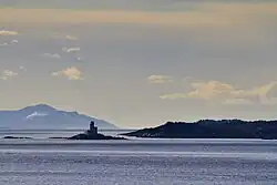 Foto einer Küstenlandschaft, im Zentrum eine kleine Insel mit einem Leuchtturm, weiter entfernt höhere Erhebungen