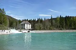 Über einem Wasserspiegel mit gletschergrünem Seewasser sieht man ein steiniges Ufer, das in Wald übergeht. In der linken Bildhälfte sieht man ein weißes Bauwerk mit hohen, schmalen Fenstern, neben dem sich schäumend Wasser über drei Stufen hinweg in den See ergießt.