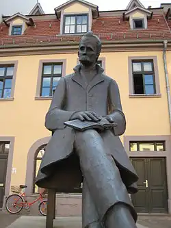 Statue von Nietzsche aus Metall, er sitzt mit übereinandergeschlagenen Beinen auf einem Stuhl und hat ein Buch auf dem Knie liegen