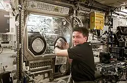 A man wearing a blue polo shirt reached into a large machine. The machine has a large windows at the front with two holes in it for access, and is full of scientific apparatus. Transient space station hardware is visible in the background.