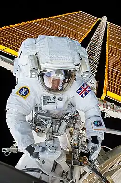 Britischer Astronaut in einem weißen Raumanzug im Weltall vor Solarpaneelen der Internationalen Raumstation