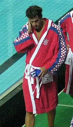 Igor Hinić trägt einen roten Bademantel mit rot-weißen Karos auf den Ärmeln. In der linken Hand hält er die Bademütze mit der Nummer 11.