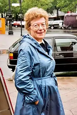 Inge Berner, schlank, groß, mit rötlichem lockigem Haar und großer Brille, lächelnd, bei einem Berlinbesuch auf dem Kurfürstendamm in einem leuchtend blauen Regenmantel vor einem Auto stehend