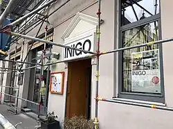 Altes Haus in Nahaufnahme mit einem Gerüst für Fassadenrenovierung davor. Über dem Eingangstor in der Mitte ist ein Schild mit der Aufrschrift „Inigo“.
