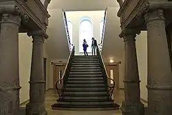 Es ist der Eingangsbereich des Paderborner Rathauses zu sehen. Mittig die Treppe, mit den ornamentalen Eisengeländern. Im Hintergrund die Rückwand des Treppenhauses mit seinen Bogenfenstern. Am Bildrand vordergründig zwei Kapitelle auf ionischen Säulen