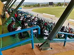 Blick von schräg oben über ein Geländer auf die Ränge des Fußballstadions. Hauptsächlich junge Männer sitzen jeweils mit Abstand auf den Plätzen. Sie tragen Gesichtsmasken und haben Prüfungsbögen auf den Knien, die sie bearbeiten. Im Hintergrund ist noch eine Ecke des Fußballplatzes und die dahinter liegende, locker bewaldete Landschaft zu erkennen.