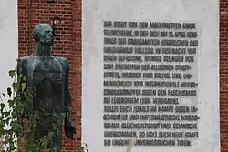Eine Bronzestatue zeigt einen abgemagerten Mann und steht vor einer weiß gestrichenen Fassade, an die mit Großbuchstaben eine fünfzehnzeilige Inschrift angebracht ist