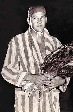 Ivo Cipci trägt auf diesem Schwarz-Weiß-Foto eine Bademütze und einen längsgestreiften Bademantel. In den Händen hält er einen mit durchsichtigem Plastik geschützten Blumenstrauß.