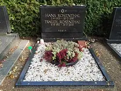 Das Bild zeigt ein Grab mit einem schwarzen Grabstein, auf dem in goldener Schrift die Namen Hans Rosenthal und Traudl Rosenthal eingraviert sind. Oberhalb der Namen ist ein Davidstern abgebildet, der die Verbundenheit zur jüdischen Religion symbolisiert. Die Inschrift lautet: "HANS ROSENTHAL 2.4.1925 - 10.2.1987 TRAUDL ROSENTHAL 6.8.1927 - 25.3.2016. "Zusätzlich gibt es eine hebräische Inschrift unter den Namen. Das Grab ist mit weißen Kieselsteinen bedeckt, die Tradition und Respekt ausdrücken. In der Mitte des Grabes befindet sich eine runde Pflanzschale, die mit grünen und roten Pflanzen geschmückt ist. Kleine Gegenstände, wie ein umgedrehter Blumentopf und ein kleiner Stein, liegen auf dem Grab. Das Grab ist von anderen Gräbern flankiert, und im Hintergrund erstreckt sich eine dichte Hecke, die den Ort ruhig und friedlich wirken lässt.