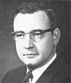 Mann im mittleren Alter von vorn ab Schulterblatt im dunklen Jackett, weißem Hemd und dunkler Krawatte in schwarzweiß.