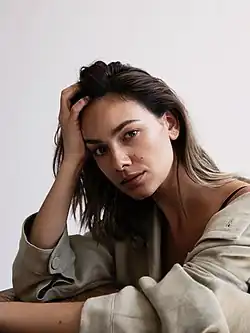 Janina Uhse (2019) mit langen Haaren, die ein helles Oberteil mit einem schwarzen Band darunter trägt. Sie stützt ihren Kopf auf eine Hand, während der andere Arm auf einer Fläche ruht. Der Hintergrund des Bildes ist schlicht und in einer hellen Farbe gehalten.