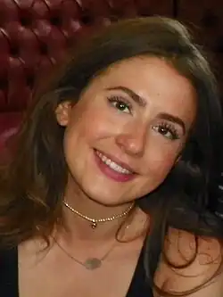 Porträtfoto von Jasmine Armfield auf dem EastEnders Meet and Greet event im BBC Elstree Centr 2016. Sie trägt gescheiteltes langes brünettes Haar. Eine Kurzkette mit kleinen runden Anhänger und einer Langkette mit einem ovalförmigen Anhänger. Schwarze dünne Träger deuten auf ein Abendkleid hin. Im Hintergrund ist ein rotes Polster einer Sitzgruppe zu erkennen.