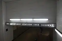 TO JAY STREET STATION IND SUBWAY. Fußgängerpassage mit Treppe, die hinab zu einer weiß gefliesten und mit Leuchtstofflampenkästen illuminierten Fußgängerpassage führt. Über dem Zugang ist ein Schriftzug, der als grauer Lichtkasten mit ausgeschnittenen Lettern über die volle Breite der Passage ausgeführt ist. Die Lettern sind weiß hinterleuchtet und bilden einen Schriftzug im Stil des Art déco.