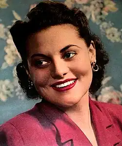 Jeanne Cagney Foto des Cover der New York Sunday News 1942. Sie trägt eine Jacke in einer weinroten Farbe. Das Haar der Person ist dunkel und stilvoll frisiert. Der Hintergrund des Bildes besteht aus floralen Mustern in Blau- und Rosatönen, die dem Bild einen eleganten und lebendigen Eindruck verleihen.
