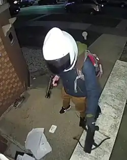 Eine Person mit Motorradhelm auf dem Kopf, die in der rechten Hand eine Schusswaffe und in der linken Hand eine Armbrust trägt