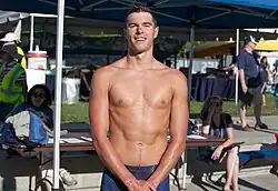 Prenot mit nacktem Oberkörper trägt eine blaue Badehose mit schwarzen Punkten. Im Hintergrund sind aufgeklappte Sonnenschirme zu sehen.