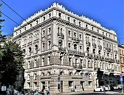 Farbige Eckansicht eines mehrgeschossigen, hellen Gebäudes mit Ornamenten an Fenstern. An manchen Fenstern sind kleine Balkone und Kopfreliefs. Das Dachgesimse besteht aus verschiedenen Skulpturen.