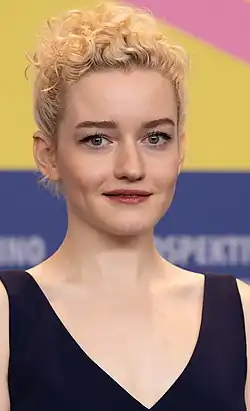Julia Garner auf der Berlinale 2020 mit lockigem, blondem Haar, das oben zu einer voluminösen Frisur gestylt ist. Die Frisur fällt leicht über die Stirn, während der Rest der Haare zu einem hohen Stil geformt ist. Sie trägt ein ärmelloses Kleid in dunkelblau mit einem tiefen V-Ausschnitt, das elegant und schlicht wirkt. Der Hintergrund ist sehr farbenfroh und besteht aus Bereichen in Gelb, Blau und Rosa, die wie abstrakte Farbflächen angeordnet sind. Im Bereich des Blaus sind teilweise Buchstaben sichtbar, die jedoch nicht vollständig lesbar sind, möglicherweise als Teil eines Schriftzugs oder Designs.