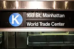 Rollbandanzeiger, weiße Schrift auf schwarzem Hintergrund. Blaues Liniensignet mit weißem Buchstaben K links, rechts zweizeilig: 168 St, Manhattan/World Trade Center