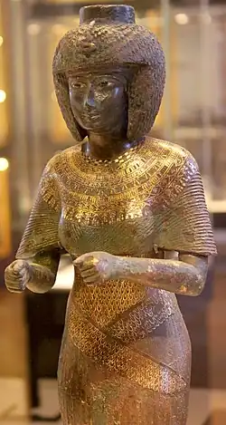 Statuette der Karomama, Hohepriesterin der 22. Dynastie, 9. Jahrhundert vor Chr., Louvre. Das enganliegende Gewand ist aus fein plissiertem Stoff, Schultern und Dekolleté sind mit einem fein gearbeiteten Schmuck aus Gold und Edelsteinen bedeckt.