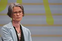 Fotoporträt einer Frau mit grauen Haaren und Brille, nur der Kopf ist zu sehen. Die Frau blickt ernst.