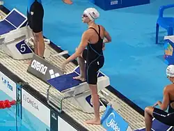 Stewart trägt einen dunkelblauen Badeanzug und eine Schwimmbrille. Auf der weißen Badekappe ist die US-Flagge und ihr Nachname zu sehen. Stewart hat das rechte Bein bereits auf dem Startblock, das linke Bein steht noch auf dem Beckenrand.