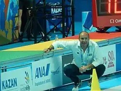 Ivica Tucak sitzt in der Hocke am Beckenrand. Er trägt ein weißes Hemd, eine blaue Hose und weiße Turnschuhe. Um den Hals baumelt seine Akkreditierung.