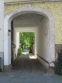 Torbogen in einer Hauserfassade mit geöffnetem Tor. Im gepflasterten Boden sind Gleise verlegt, die vom Gehweg in Richtung Hinterhof wegführen