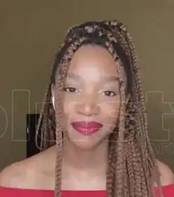 Khosi Ngema mit langen, geflochtenen Haaren, die ein rotes schulterfreies Oberteil trägt und schaut lächelnd in die Kamera. Der Hintergrund ist schlicht und scheint sich in einem Innenraum zu befinden.