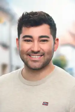 Ein lächelnder Mann mit kurzen dunklen Haaren und Bart, der einen hellen Pullover trägt und im Freien steht, wobei die Gebäude im Hintergrund verschwimmen.