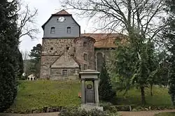 Blick aus Dorfrichtung mit vorgelagertem Kriegerdenkmal, in der Mitte des Kirchengebäudes der Treppenturm